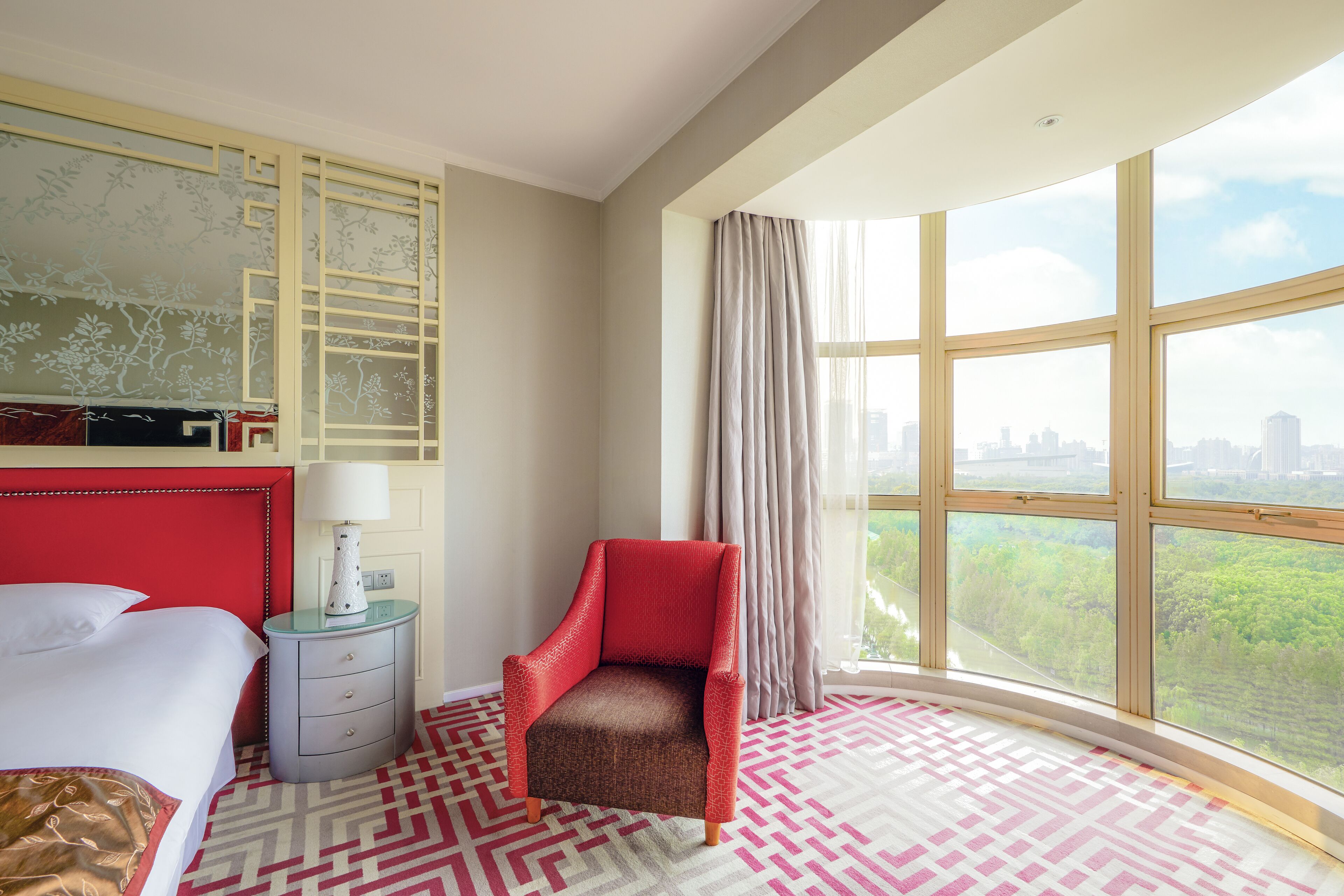 Una habitación de hotel con un ventanal grande, un sillón rojo, una cama y una mesita de noche con una lámpara.