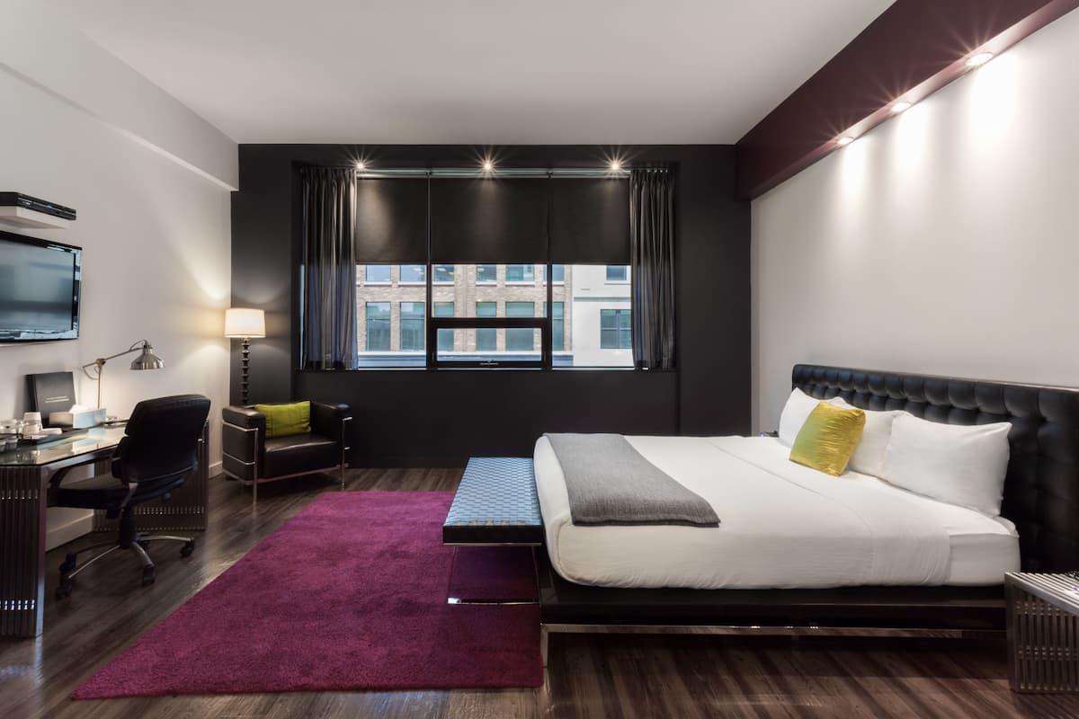 Habitación de hotel moderna con una cama grande, un escritorio, una silla, un televisor y un ventanal con cortinas.