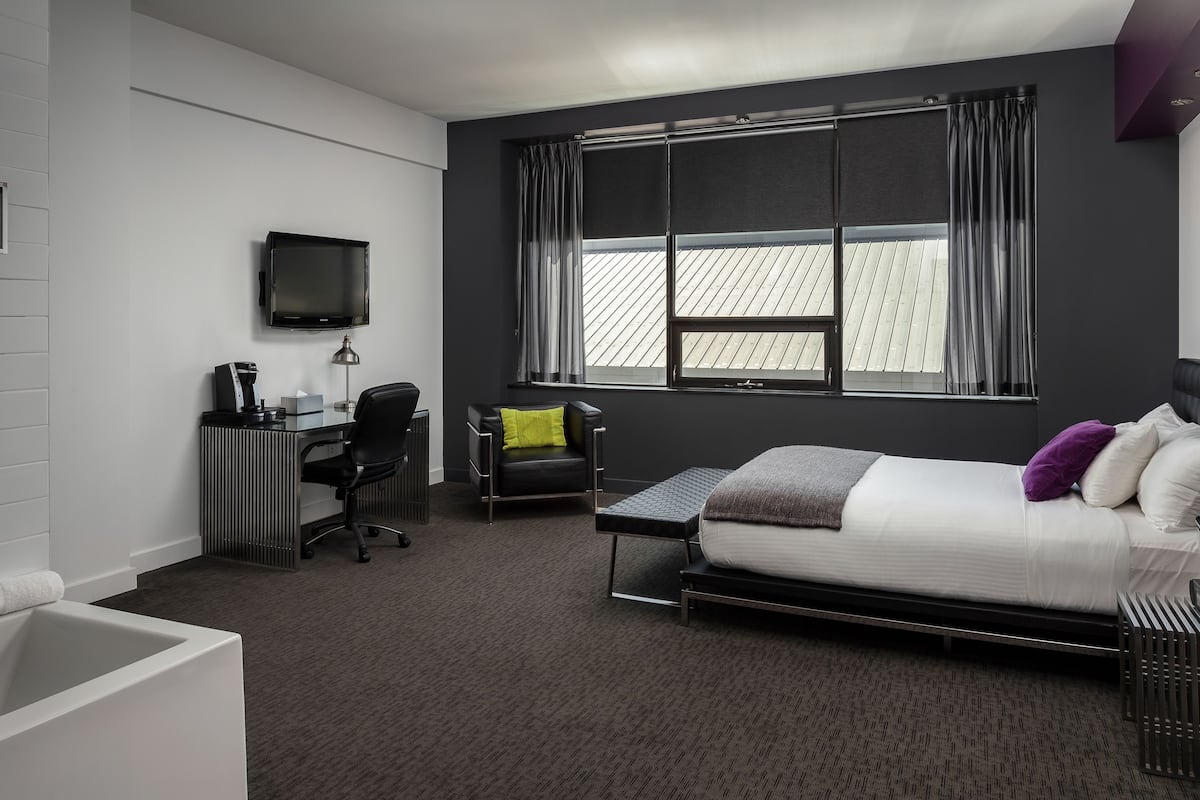 Habitación de hotel moderna con cama, escritorio, silla, televisor y un amplio ventanal con cortinas.