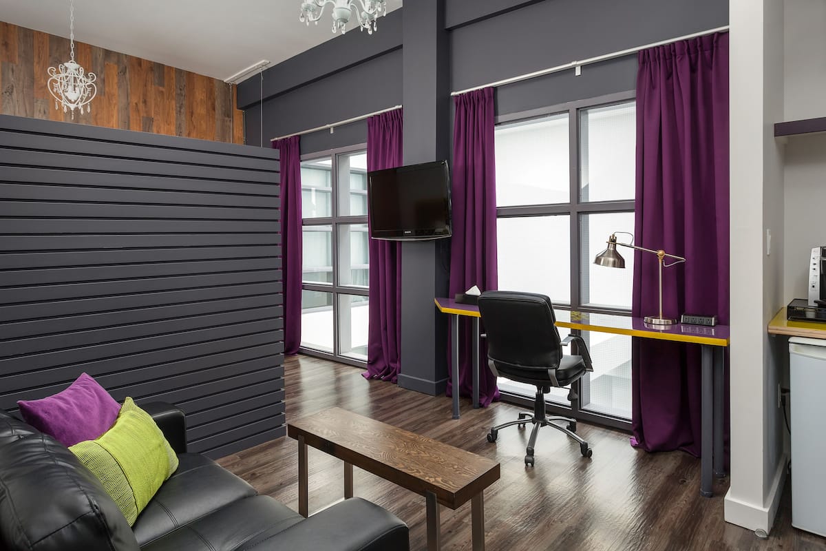 Una habitación moderna con una pared de madera, un sofá de cuero negro, una mesa de centro de madera, un escritorio con una computadora y cortinas moradas.