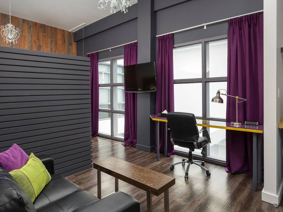 Una habitación moderna con una pared de madera, un sofá de cuero negro, una mesa de centro de madera, un escritorio con una computadora y cortinas moradas.