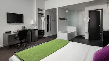 Une chambre d’hôtel comprenant un lit, un bureau, une chaise, une télévision, une salle de bain avec un lavabo et un miroir, et un peignoir accroché au mur.