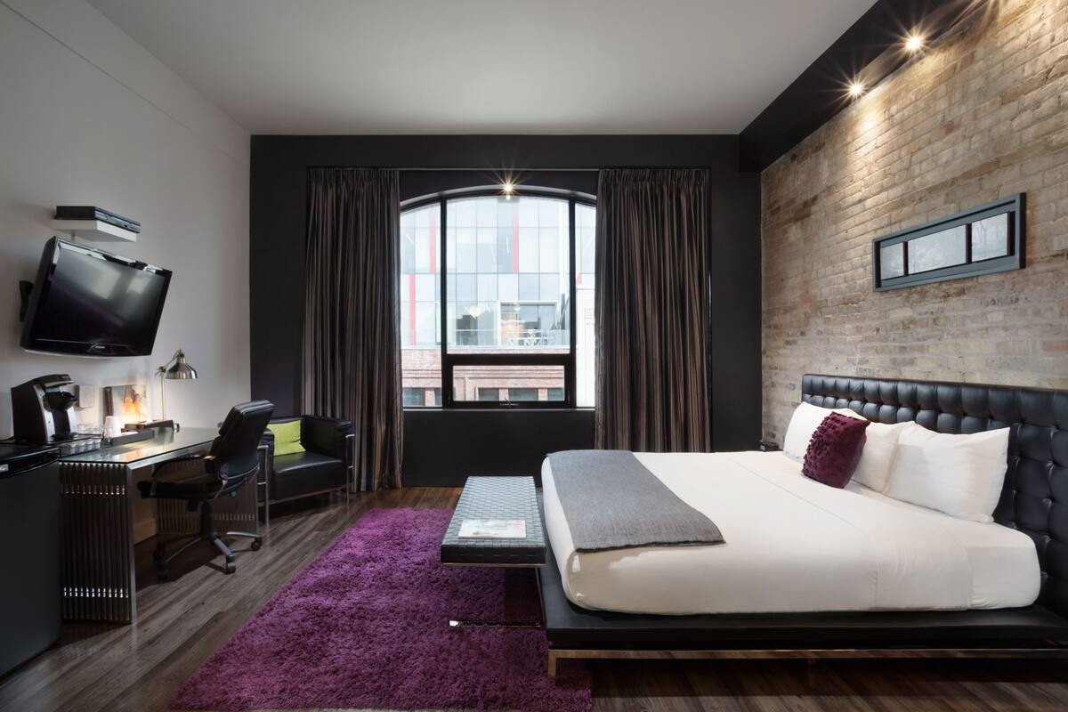 Una habitación de hotel moderna con una cama grande, un escritorio con una computadora, un televisor y vistas a la ciudad a través de un ventanal amplio.