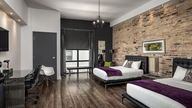 Habitació d'hotel moderna amb paret de maó, dos llits, una escriptori i un finestral gran.