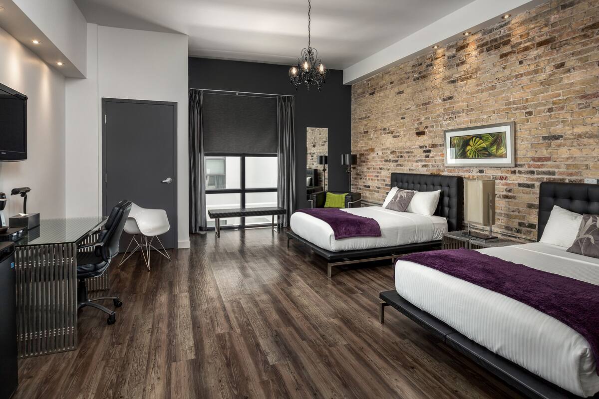 Una habitación de hotel moderna con pared de ladrillo, dos camas, un escritorio y un ventanal grande.