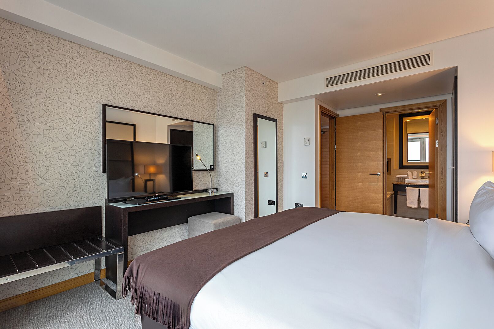 A hotel room with a bed, a desk with a mirror, a chair, and a bathroom visible through an open door.