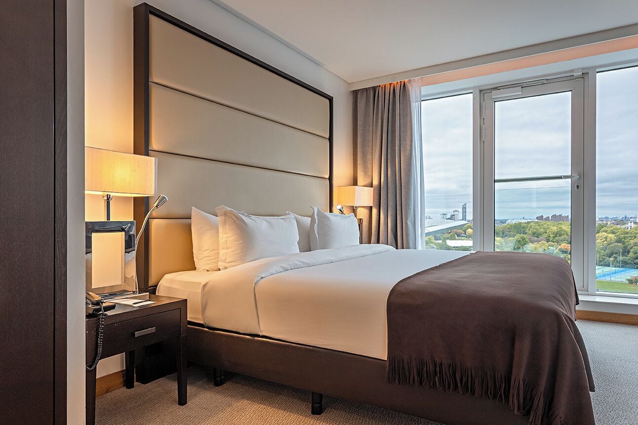 Una habitación de hotel con una cama grande, dos mesitas de noche con lámparas y vistas a la ciudad a través de amplios ventanales.