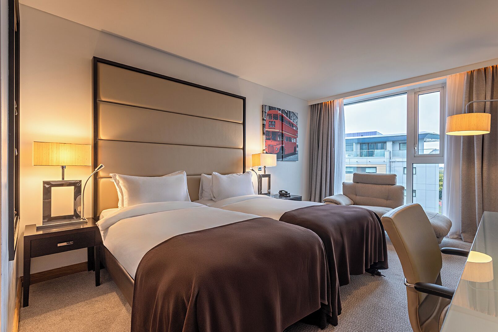 A hotel room with two beds, a large window, a chair, and a lamp.