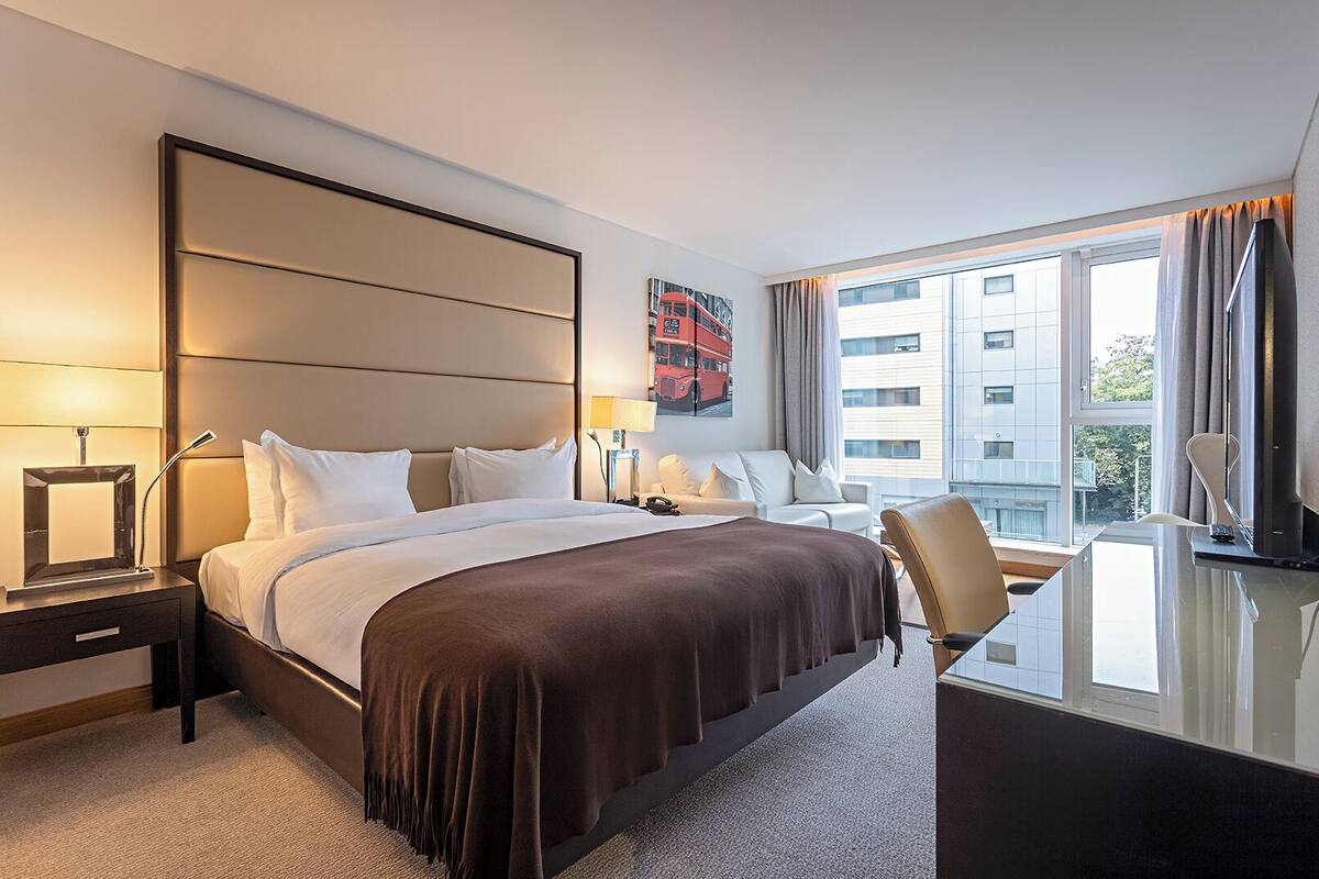 A hotel room with a large bed, bedside tables, a desk, a chair, and a window with a view of buildings.
