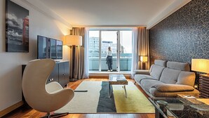 A modern living room with a sofa, armchair, TV, and a balcony with a city view. - Pestana Chelsea Bridge Hotel & SPA (London)