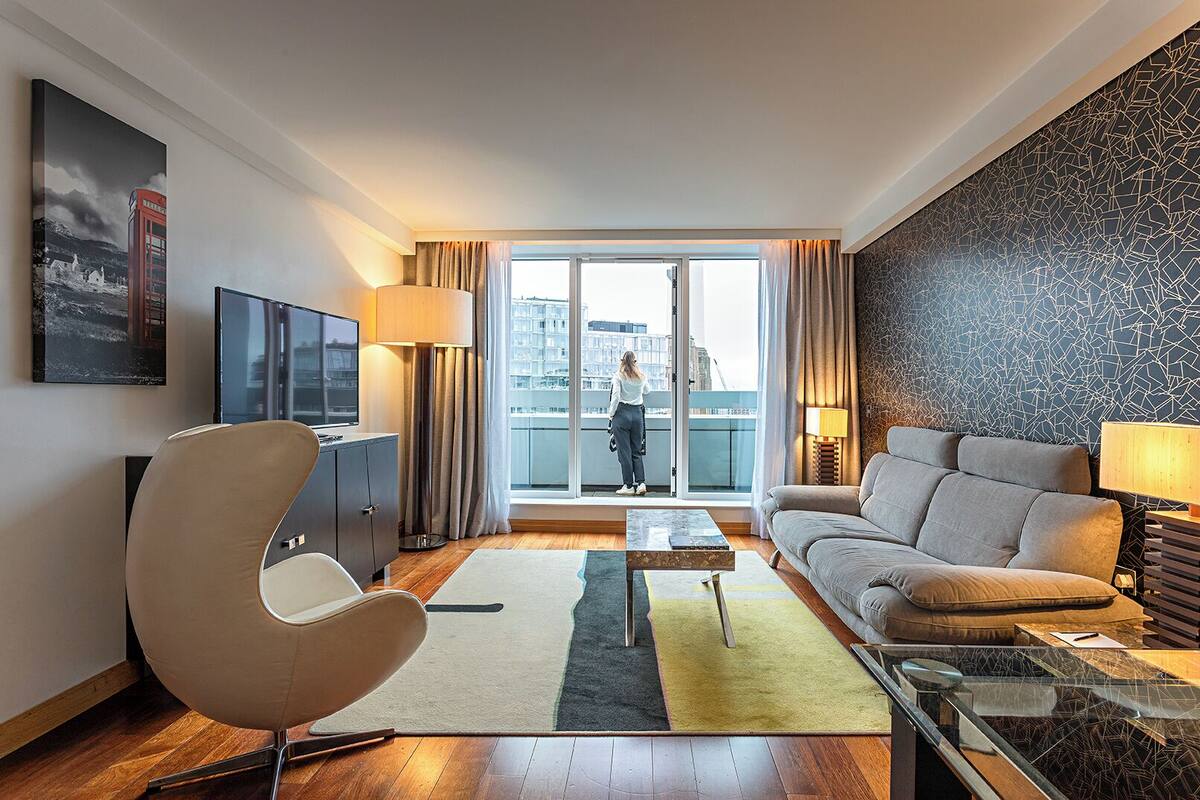 A modern living room with a sofa, armchair, TV, and a balcony with a city view.