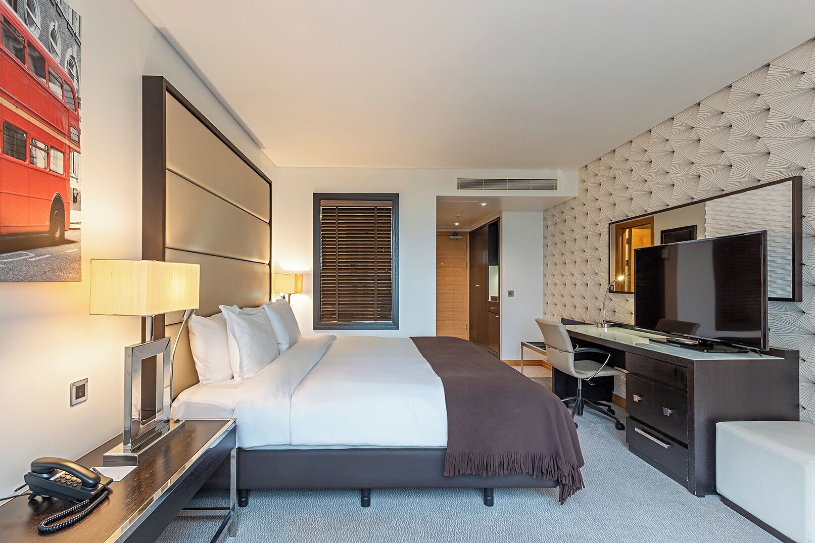 Une chambre d’hôtel équipée d’un lit, d’un bureau, d’une chaise, d’une télévision et d’une grande fenêtre avec des stores.