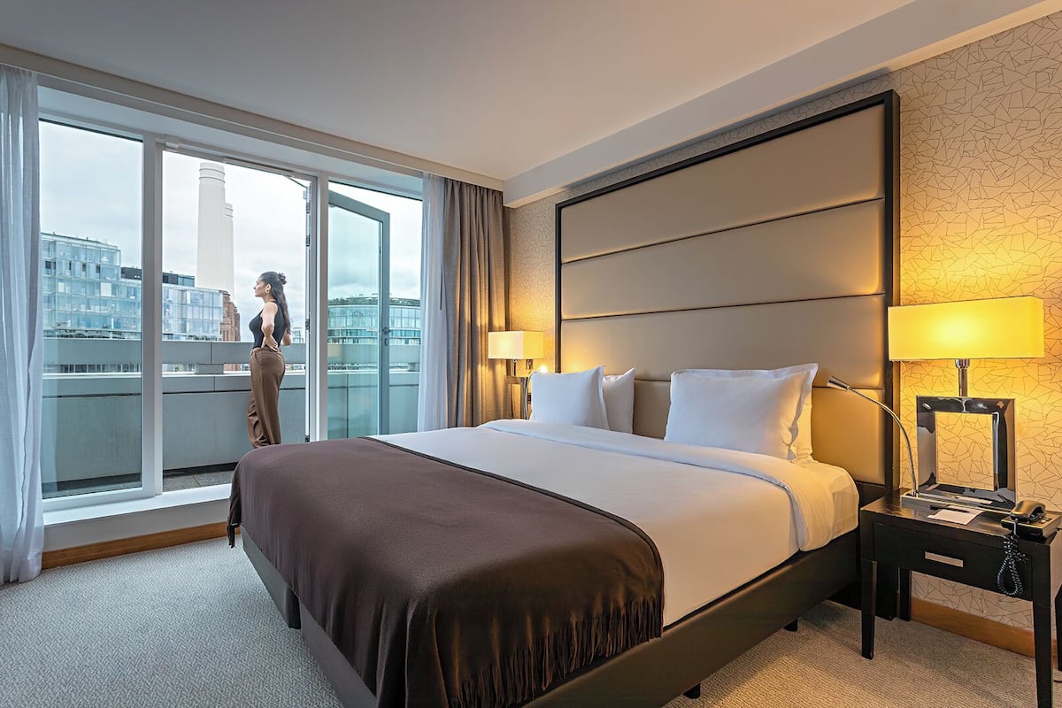 A hotel room with a large bed, two bedside lamps, a nightstand, and a view of a cityscape through a large window.