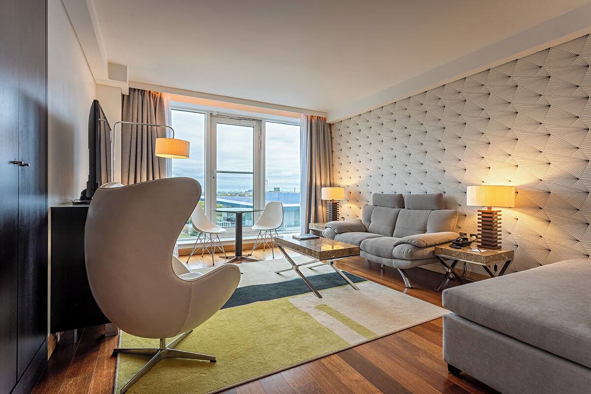 A modern hotel room with a sofa, armchair, dining table, and a view through large windows.