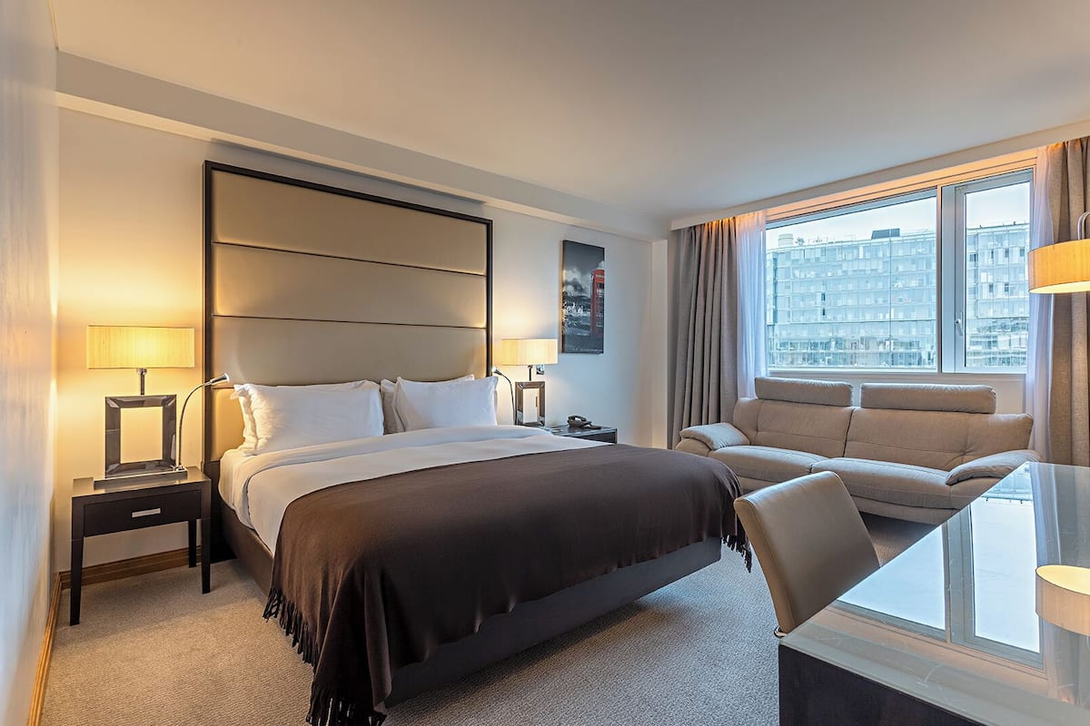 A hotel room with a bed, a desk, a chair, a nightstand, and a window with a city view.