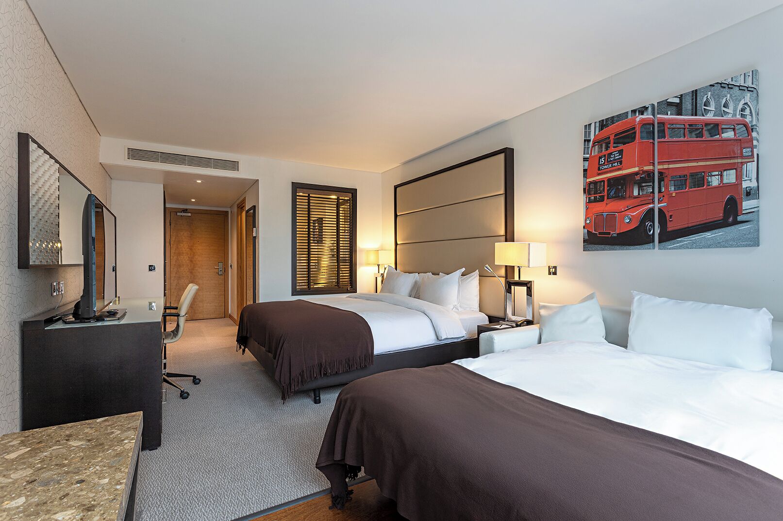 Een hotelkamer met twee bedden, een bureau, een stoel, een televisie, een lamp en een schilderij van een rode dubbeldekkerbus.