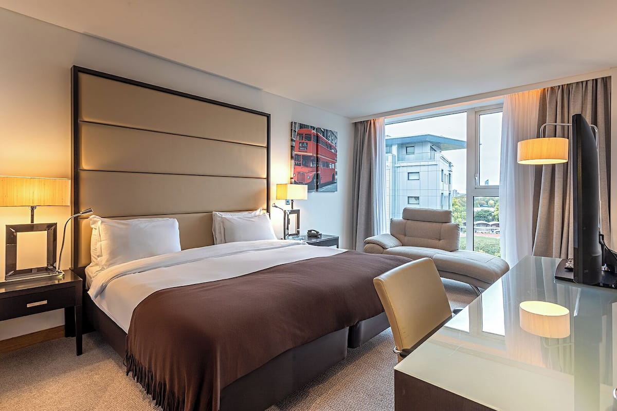 A hotel room with a bed, bedside tables, a desk, a chair, a lamp, and a window with a view of buildings.