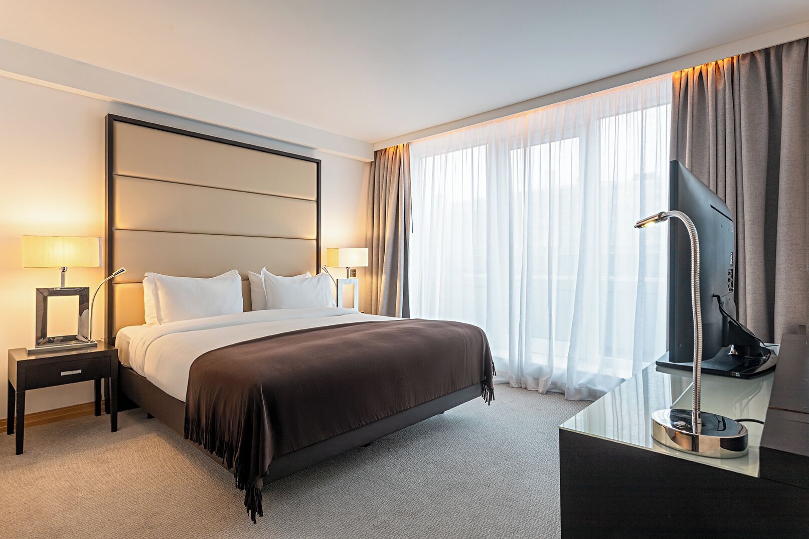 A hotel room with a large bed, a flat-screen TV, a desk with a lamp, and sheer curtains.