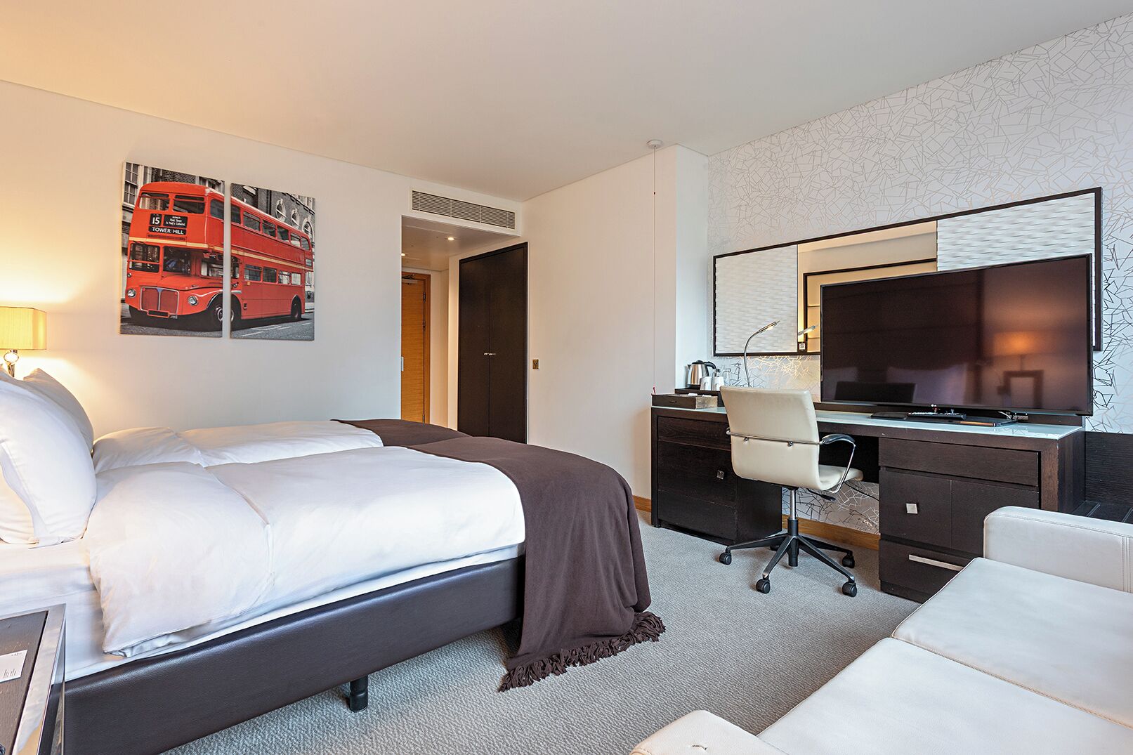 Habitación de hotel con cama doble, un escritorio con silla, televisor y una obra de arte con un autobús de dos pisos rojo en la pared.