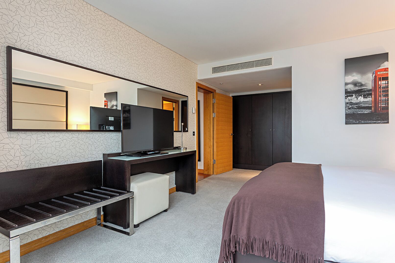 A hotel room with a bed, a desk with a TV, a wardrobe, and a door leading to another room.
