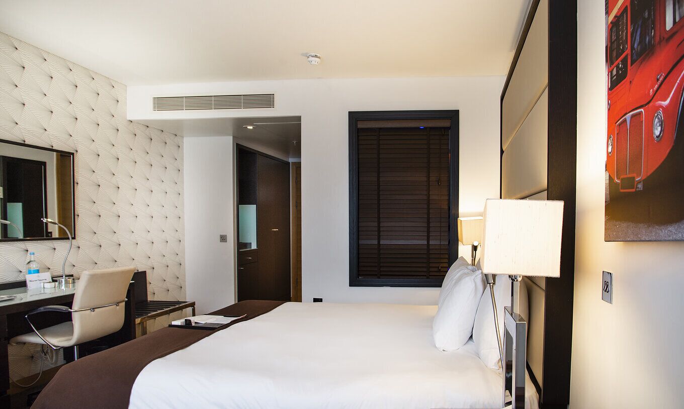 A hotel room with a bed, a desk with a chair, a mirror, a window with blinds, and a wall-mounted TV.
