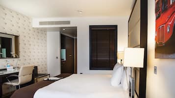 A hotel room with a bed, a desk with a chair, a mirror, a window with blinds, and a wall-mounted TV.