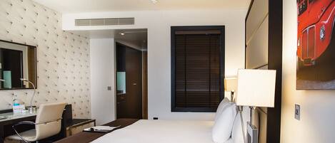 A hotel room with a bed, a desk with a chair, a mirror, a window with blinds, and a wall-mounted TV.