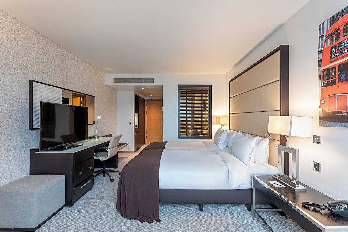 A hotel room with a bed, desk, TV, and a large window with blinds.