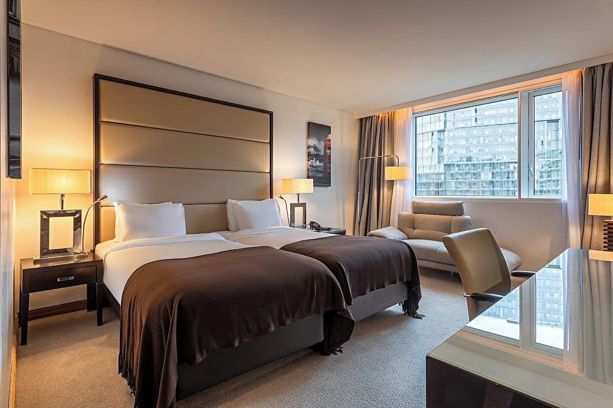 A hotel room with two beds, a sofa, a desk, and a large window with a city view.
