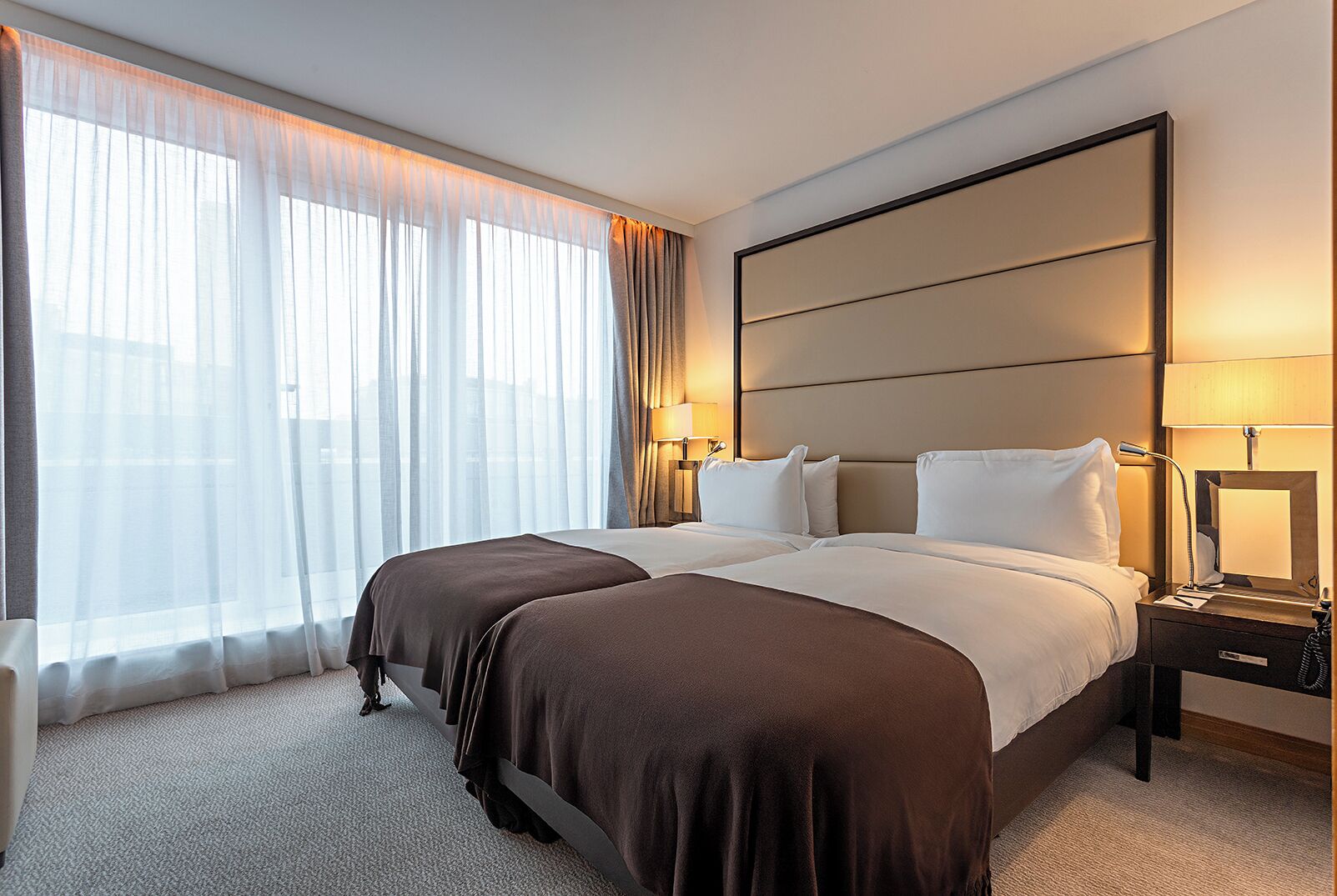 A hotel room with a large bed, two bedside tables, a nightstand, a window with sheer curtains, and a wall-mounted headboard.