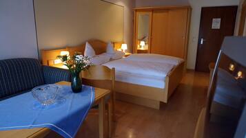 Quarto de hotel com cama, mesa de cabeceira, um vaso com flores, toalha de mesa azul, uma cadeira e porta de madeira.