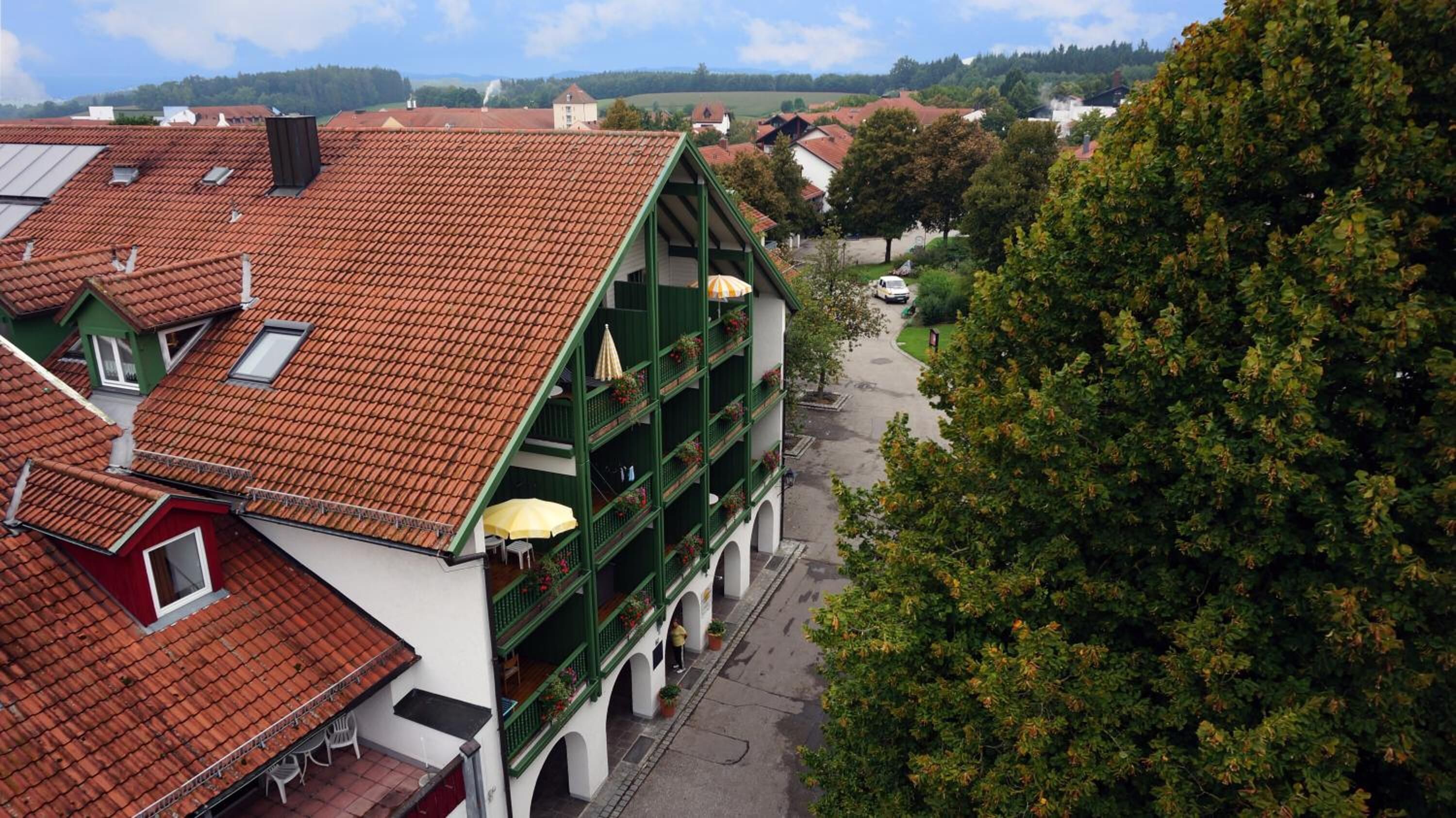 Um edifício com telhado de telhas vermelhas, várias varandas com grades verdes e vista para uma rua arborizada.