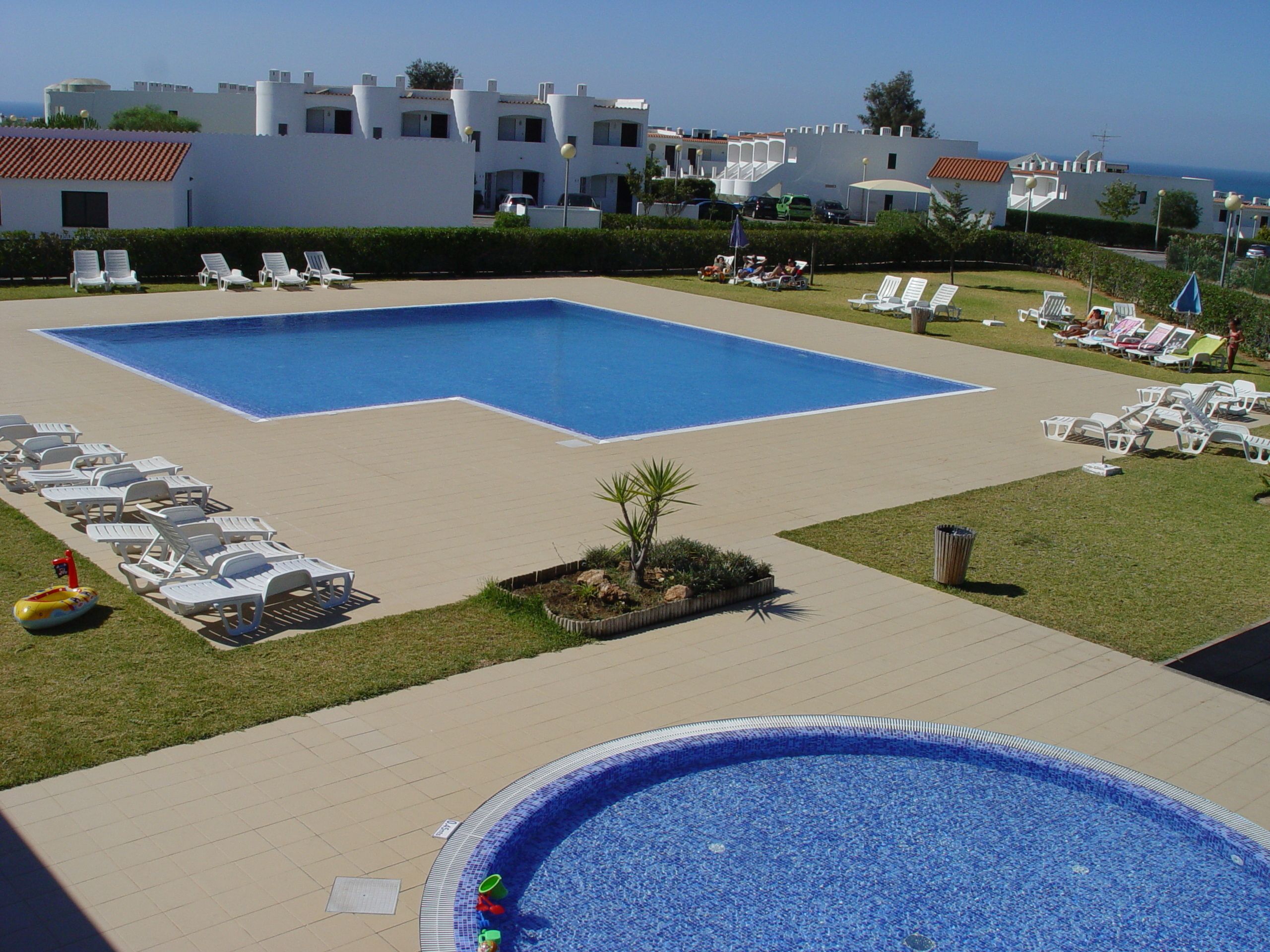 Une grande piscine rectangulaire, avec une piscine circulaire plus petite en premier plan, entourée de chaises longues blanches et d’une pelouse bien entretenue.