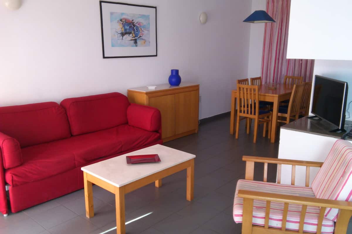 Ein Wohnzimmer mit einem roten Sofa, einem Couchtisch aus Holz, einem gestreiften Sofa, einem Esstisch mit Stühlen, einem Fernseher, einer Vase und einem Bild an der Wand.