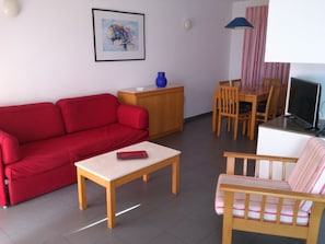 Een woonkamer met een rode bank, een houten salontafel, een gestreepte bank, een eettafel met stoelen, een televisie, een vaas en een schilderij aan de muur.