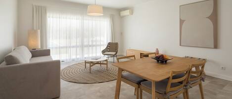 Ein modernes Wohnzimmer mit einer Couch, einem Esstisch mit Stühlen, einem runden Teppich und einem großen Fenster mit transparenten Vorhängen.
