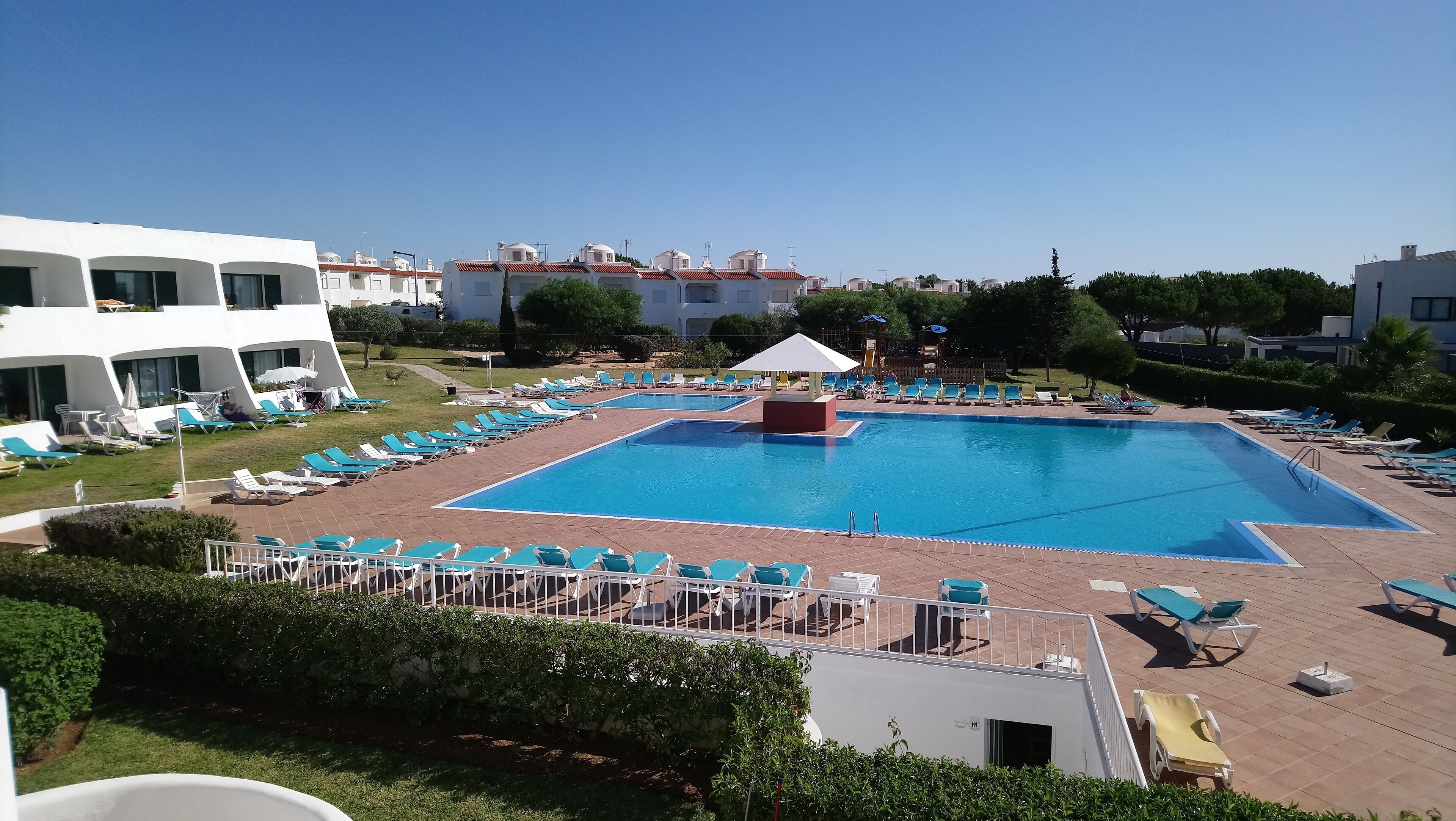 Una grande piscina con numerosi lettini prendisole e un piccolo padiglione, circondata da prati curati e edifici.