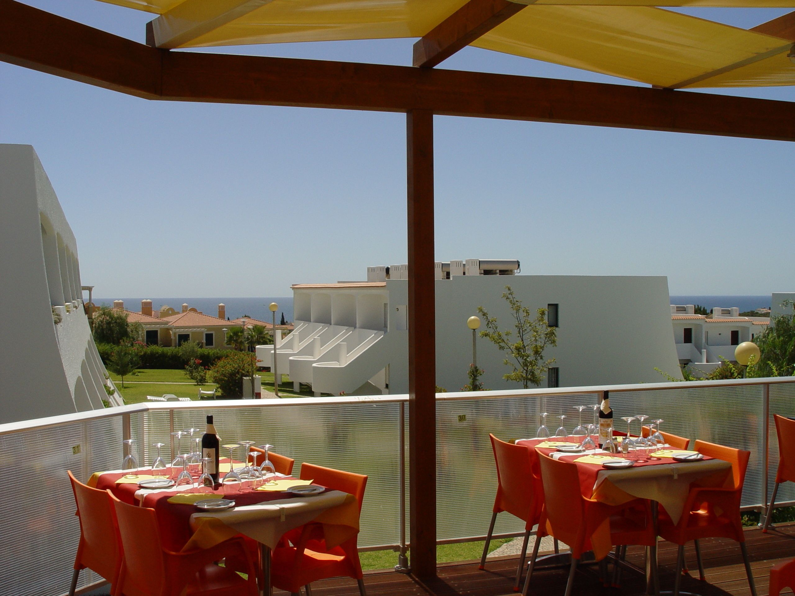 A terrace with tables set for dining, overlooking a coastal area with buildings and the sea.