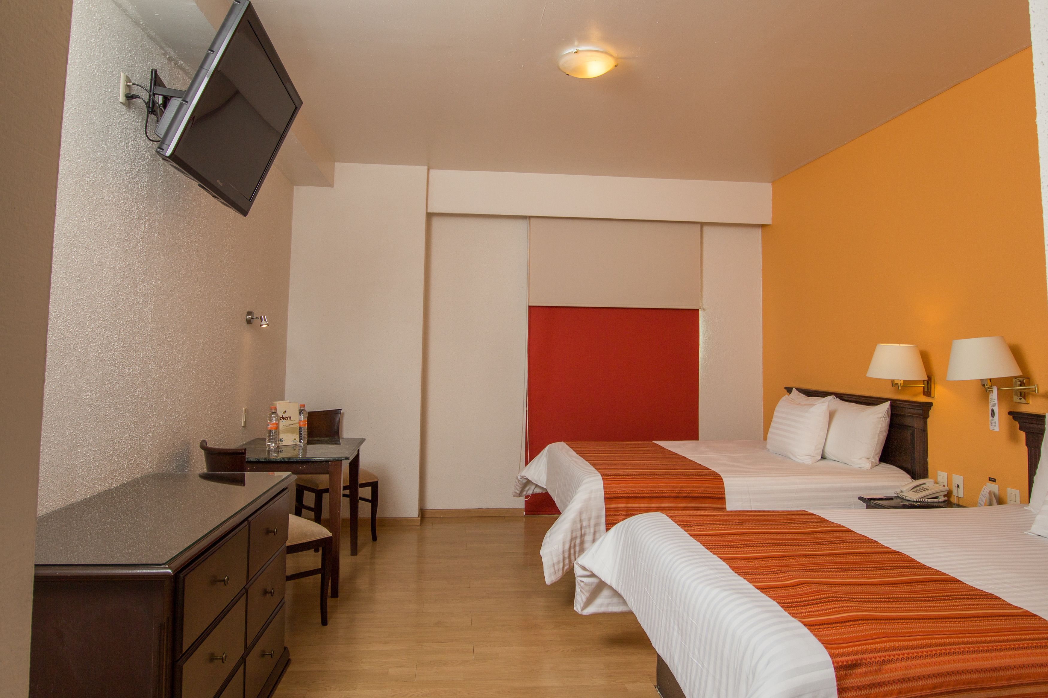A hotel room with two beds, a desk with a chair, a television mounted on the wall, and a red accent wall.