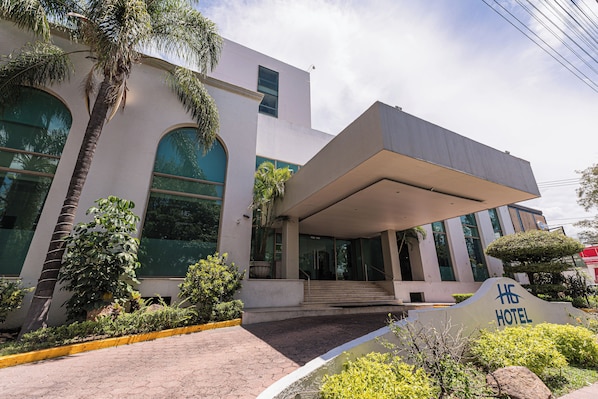 A modern hotel entrance with a covered walkway, palm trees, and a sign indicating the hotel name.