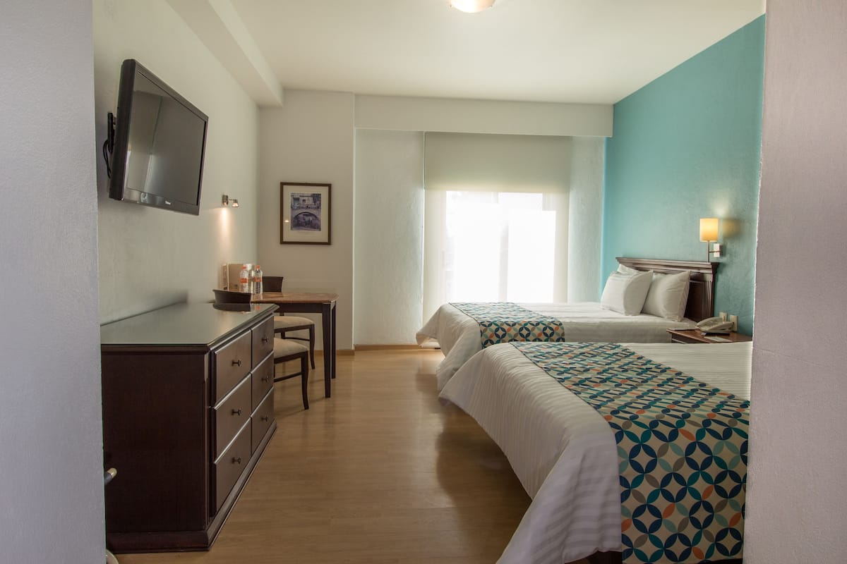 A hotel room with two beds, a desk, a TV, and a window with curtains.