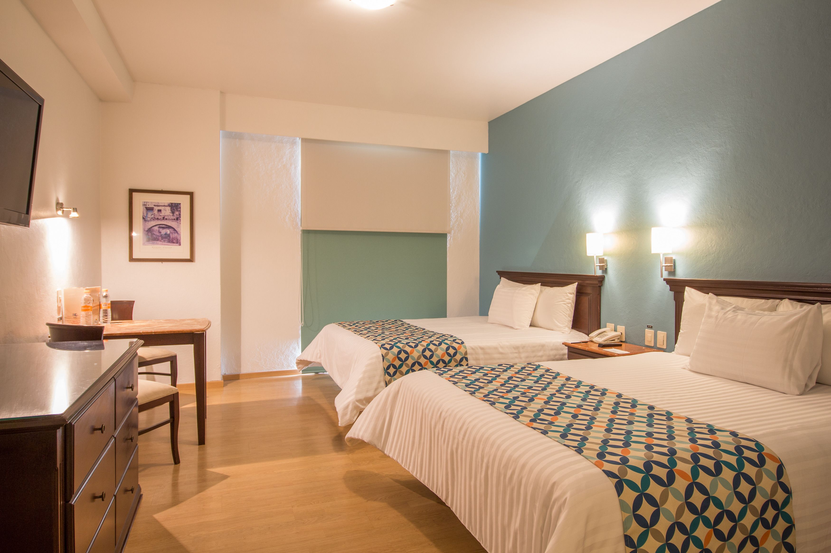 A hotel room with two beds, a desk, and a chair. There are two bedside tables with lamps.