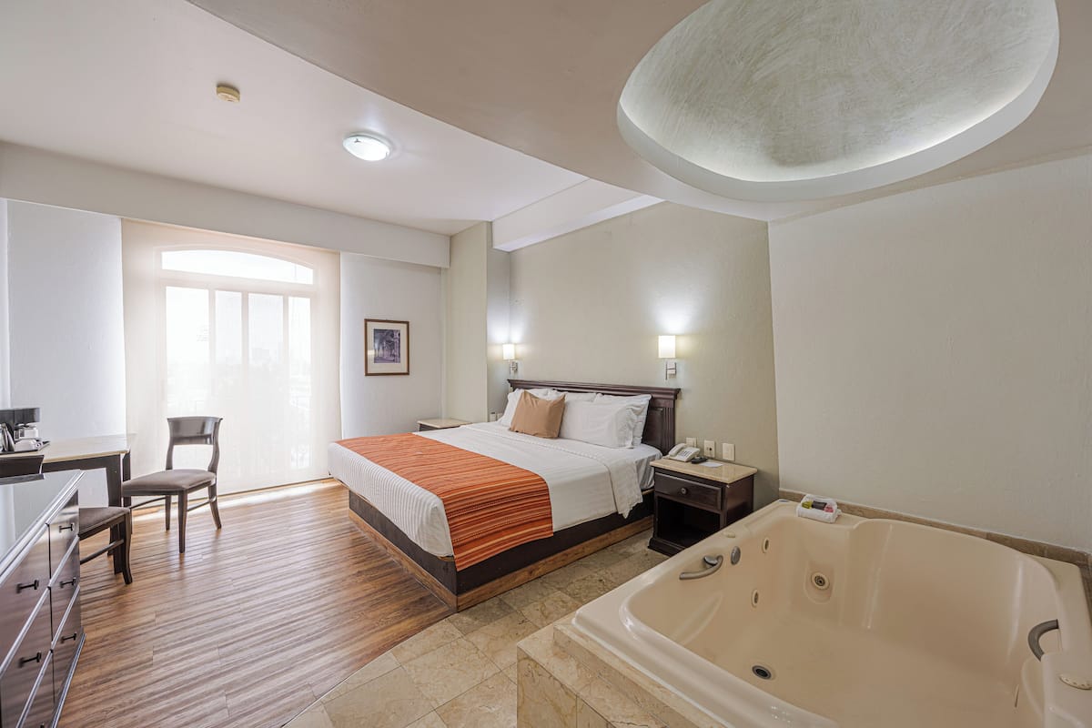 A hotel room with a wooden floor, a large bed with a white and orange bedspread, a nightstand with a lamp, a desk with a chair, and a large bathtub.