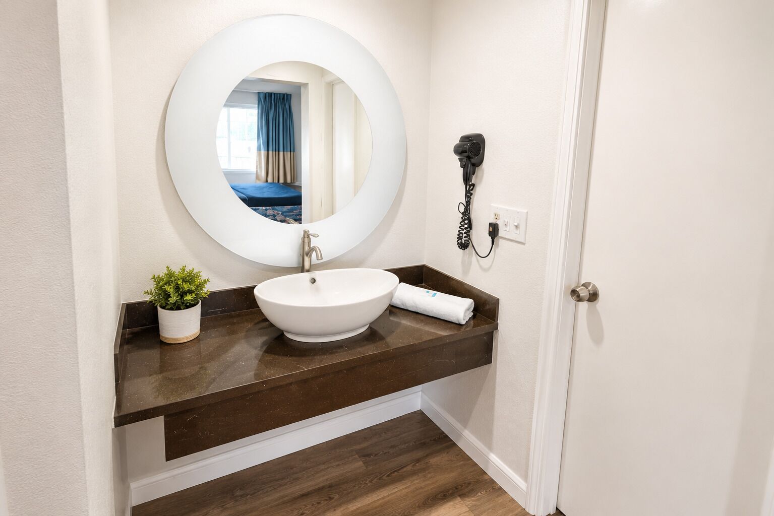 A bathroom with a round mirror, a white sink, a towel, a hairdryer, and a small potted plant.