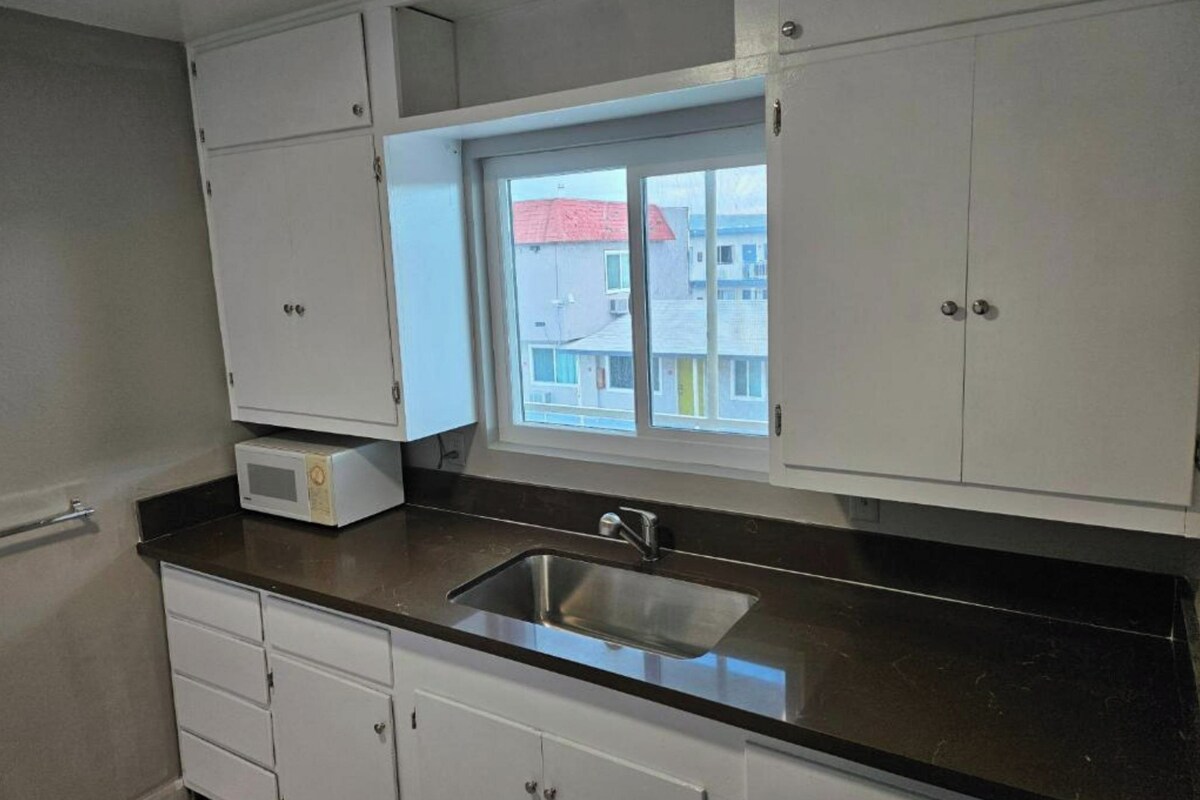 A kitchen with white cabinets, a black countertop, a stainless steel sink, and a window with a view of buildings.