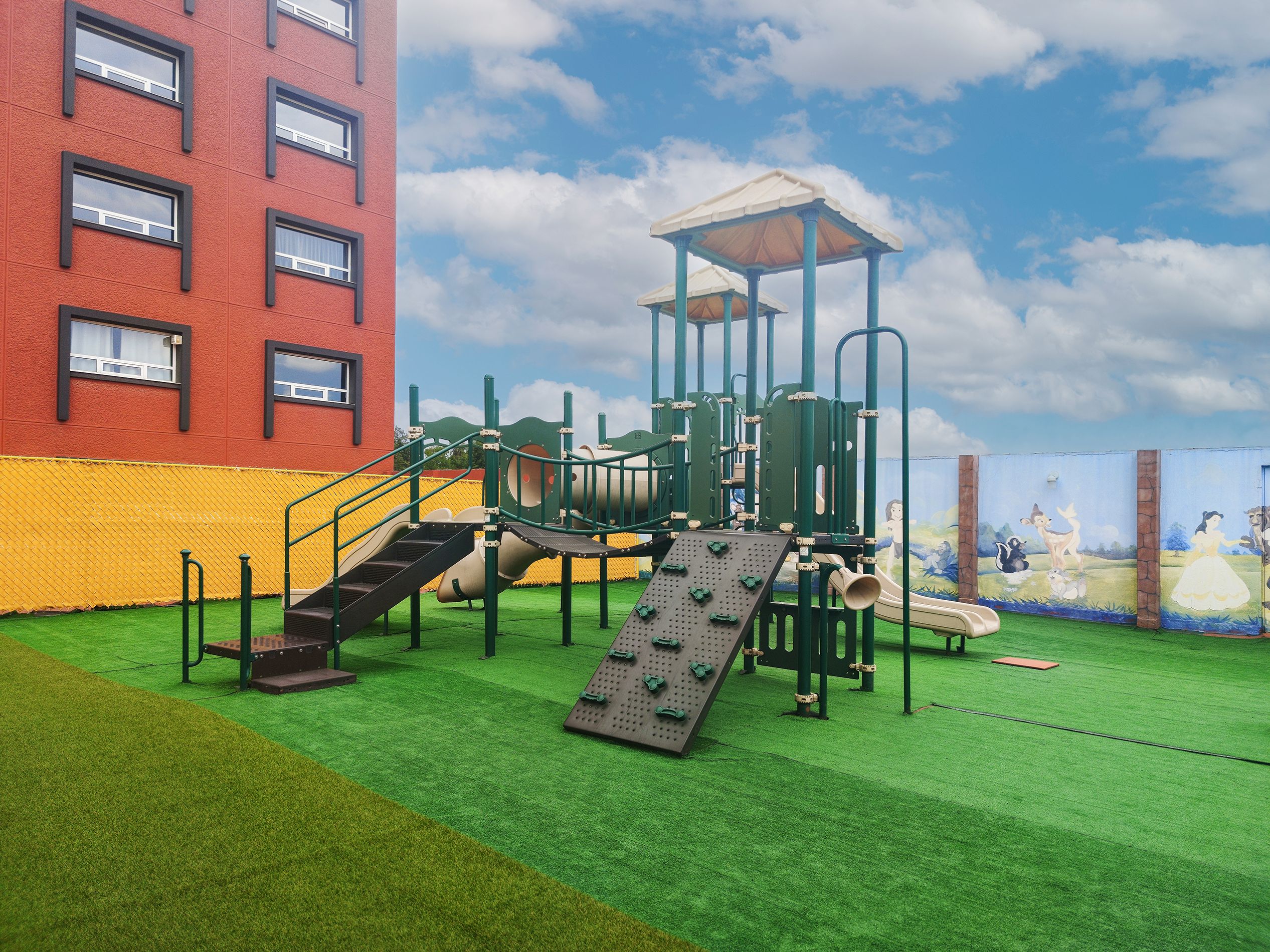 Un parque infantil con estructuras para escalar y toboganes, junto a un edificio con numerosas ventanas.