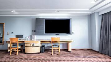A hotel room with a large flat-screen TV mounted on the wall, a wooden desk with two chairs, a small trash bin, and a patterned floor.
