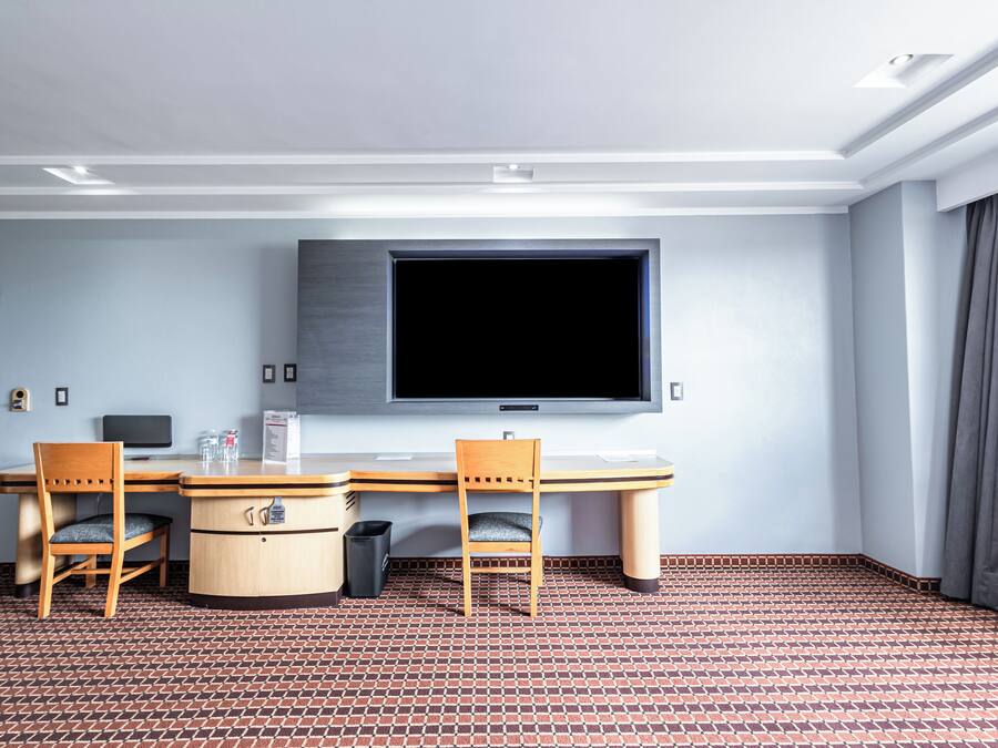 A hotel room with a large flat-screen TV mounted on the wall, a wooden desk with two chairs, a small trash bin, and a patterned floor.