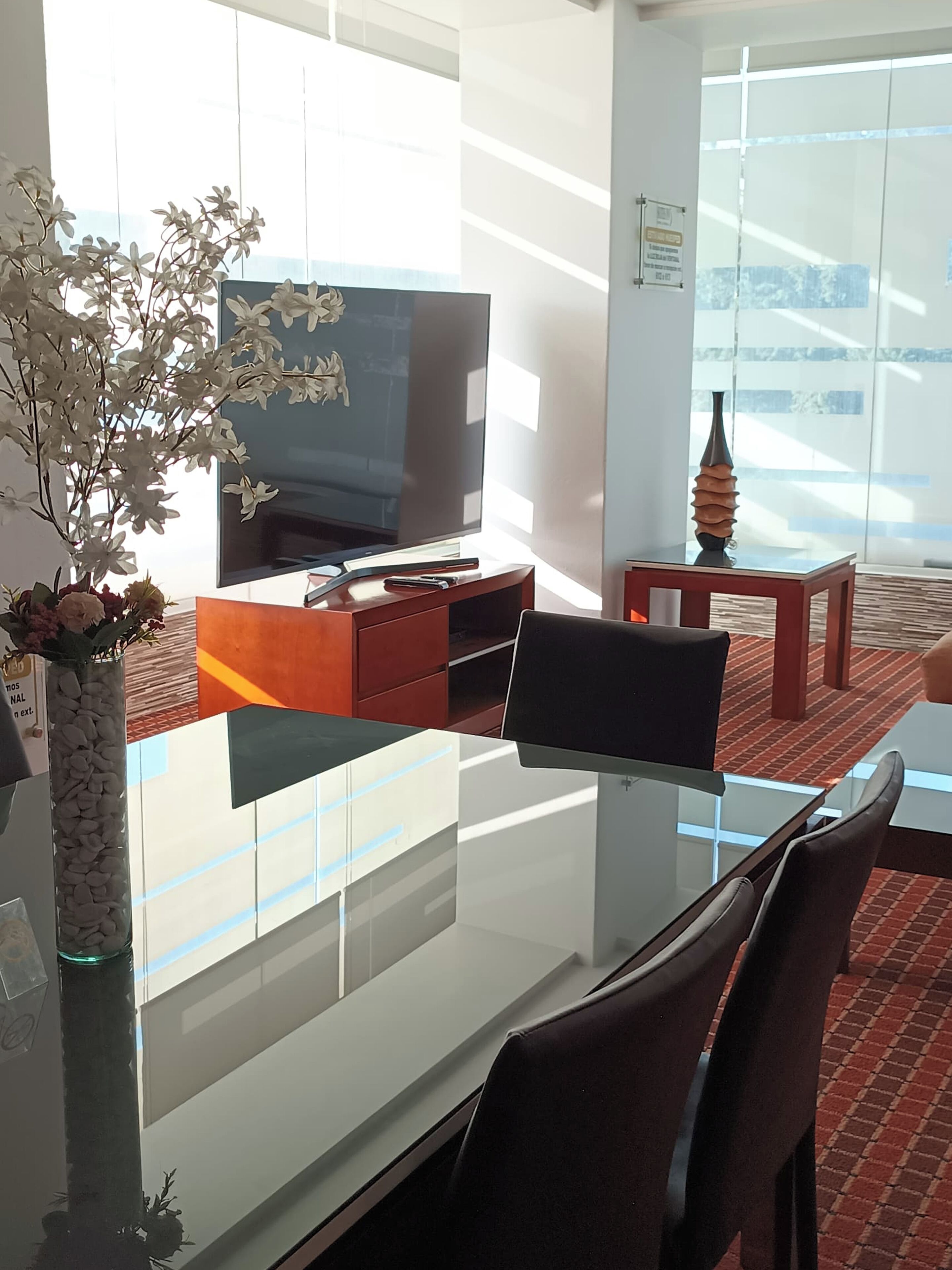 A modern living room with a glass dining table, a flat-screen TV, and a decorative vase.