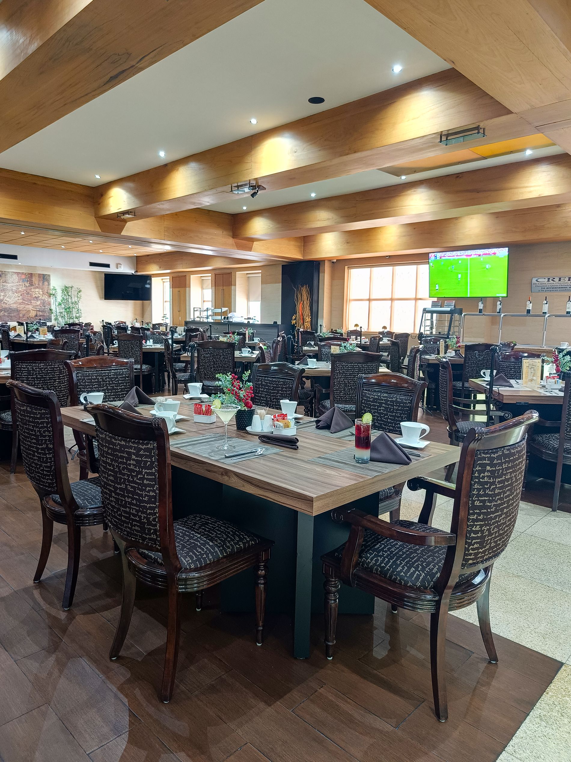 A spacious dining area with multiple tables and chairs, a flat-screen TV mounted on the wall, and a wooden ceiling.
